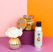 Three harvest pumpkin-themed products—a scented candle with lid, a silver fragrance spray, and the Bridgewater Candle Company Harvest Pumpkin Mercury Flower Diffuser—are arranged on a purple block against a pink and orange background. Enjoy the inviting aroma of pumpkin spice fragrance as you bring seasonal charm to any space.