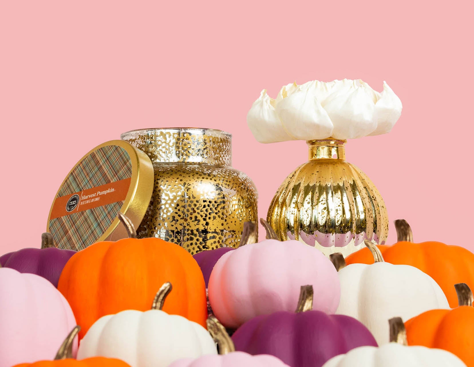 A Bridgewater Candle Company Harvest Pumpkin Mercury Flower Diffuser in a gold jar is displayed alongside a gold vase with a white flower, surrounded by small pumpkins in orange, pink, purple, and white against a plain pink background.