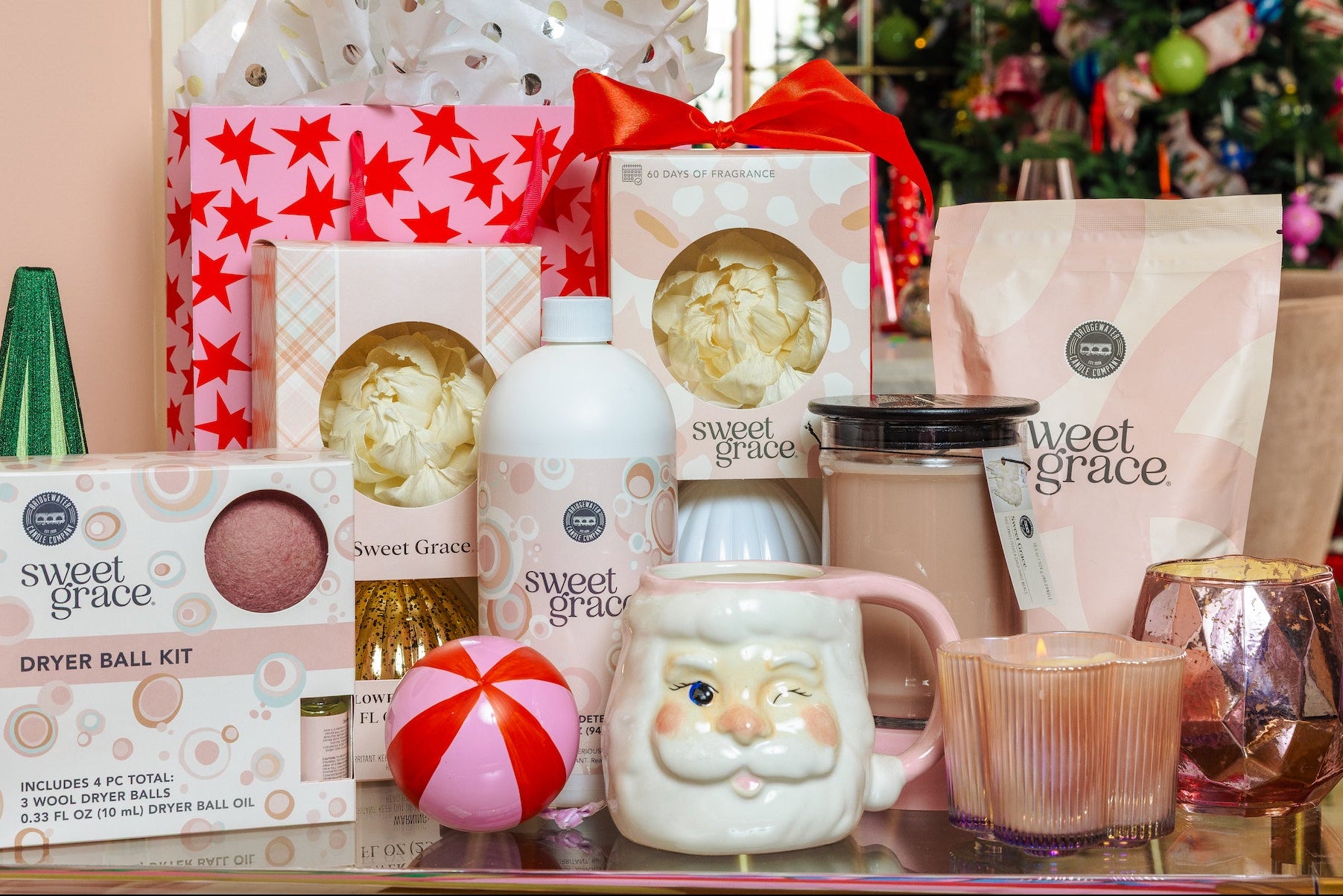 A collection of Sweet Grace brand holiday gift items arranged on a glass table in front of a decorated Christmas tree. The products include a dryer ball kit, a soap, a hot chocolate mix bag, a mug, and candles, with matching pink and white gift packaging.