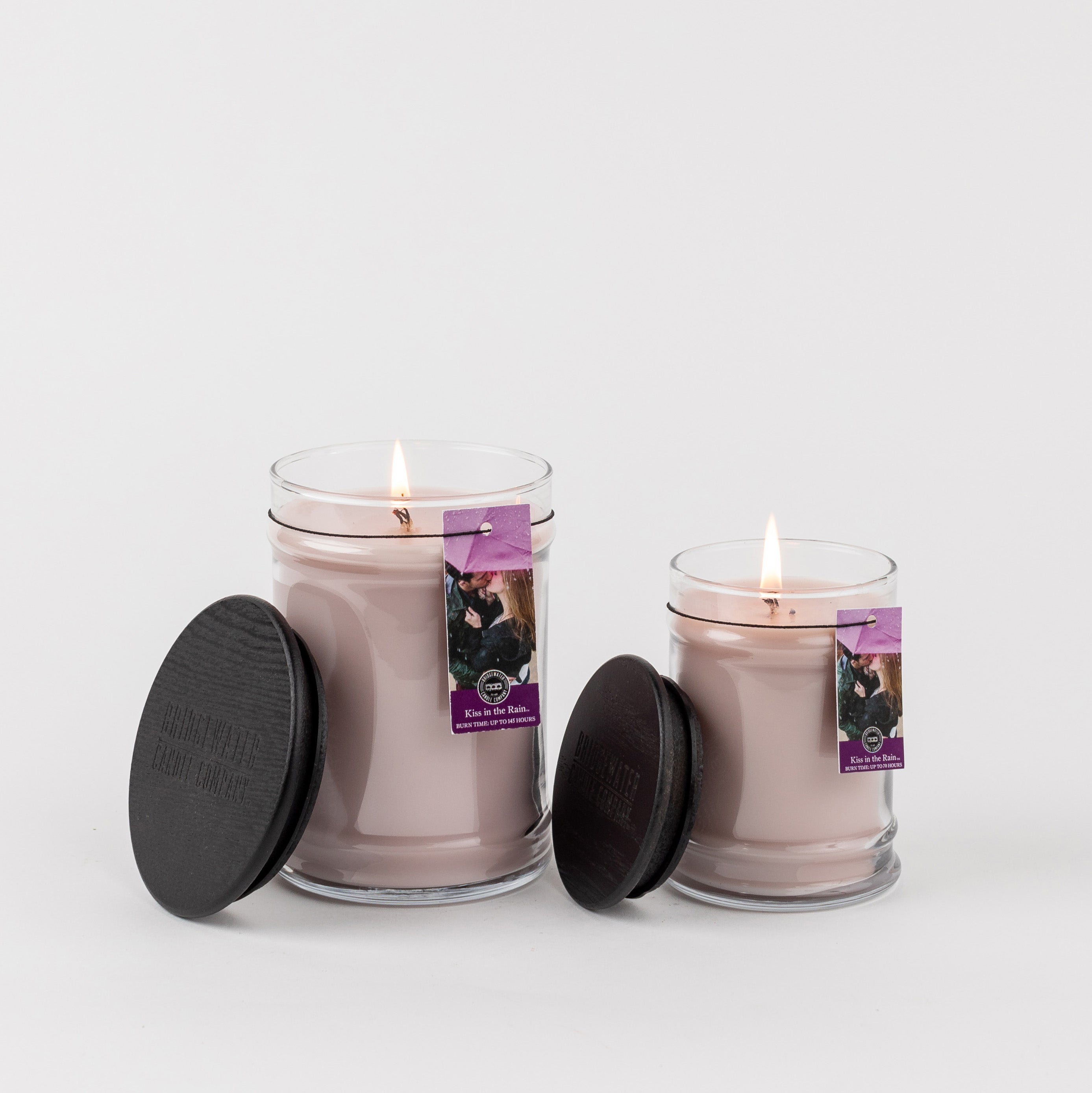 Two lit Kiss In The Rain 8oz jar candles by Bridgewater Candle Company, in glass jars with black lids and purple labels showing the artwork and text, are displayed on a plain white background.