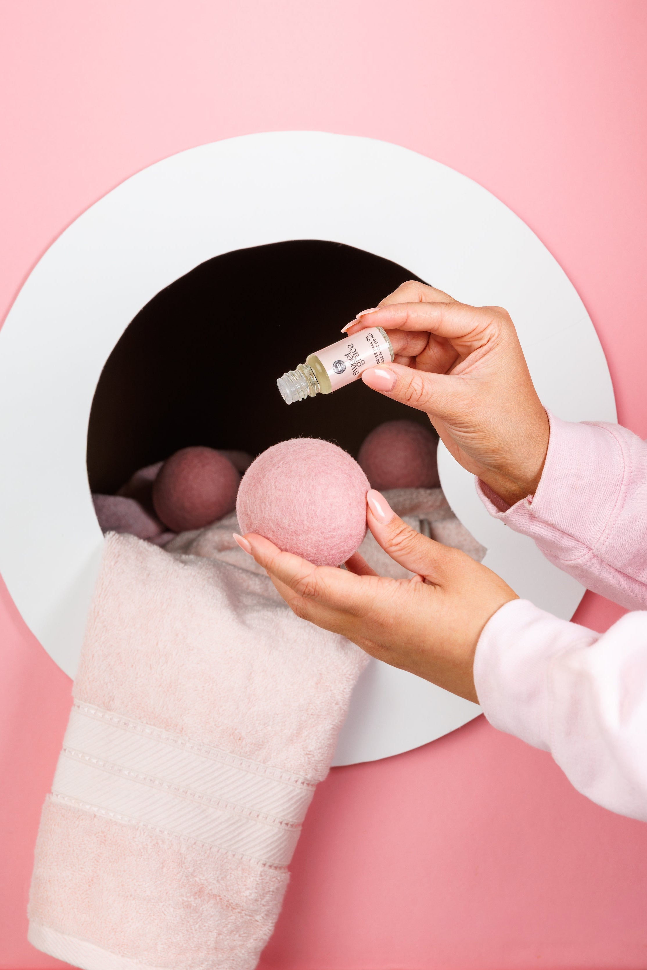 A person adds essential oil to a pink dryer ball, with other pink balls and a soft pink towel inside a dryer against a pink background.