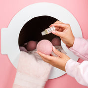 A person adds Sweet Grace Dryer Ball Kit oil by Bridgewater Candle onto a pink wool dryer ball before a white laundry machine. A pink towel and more fragranced balls are inside, all against a pink background.