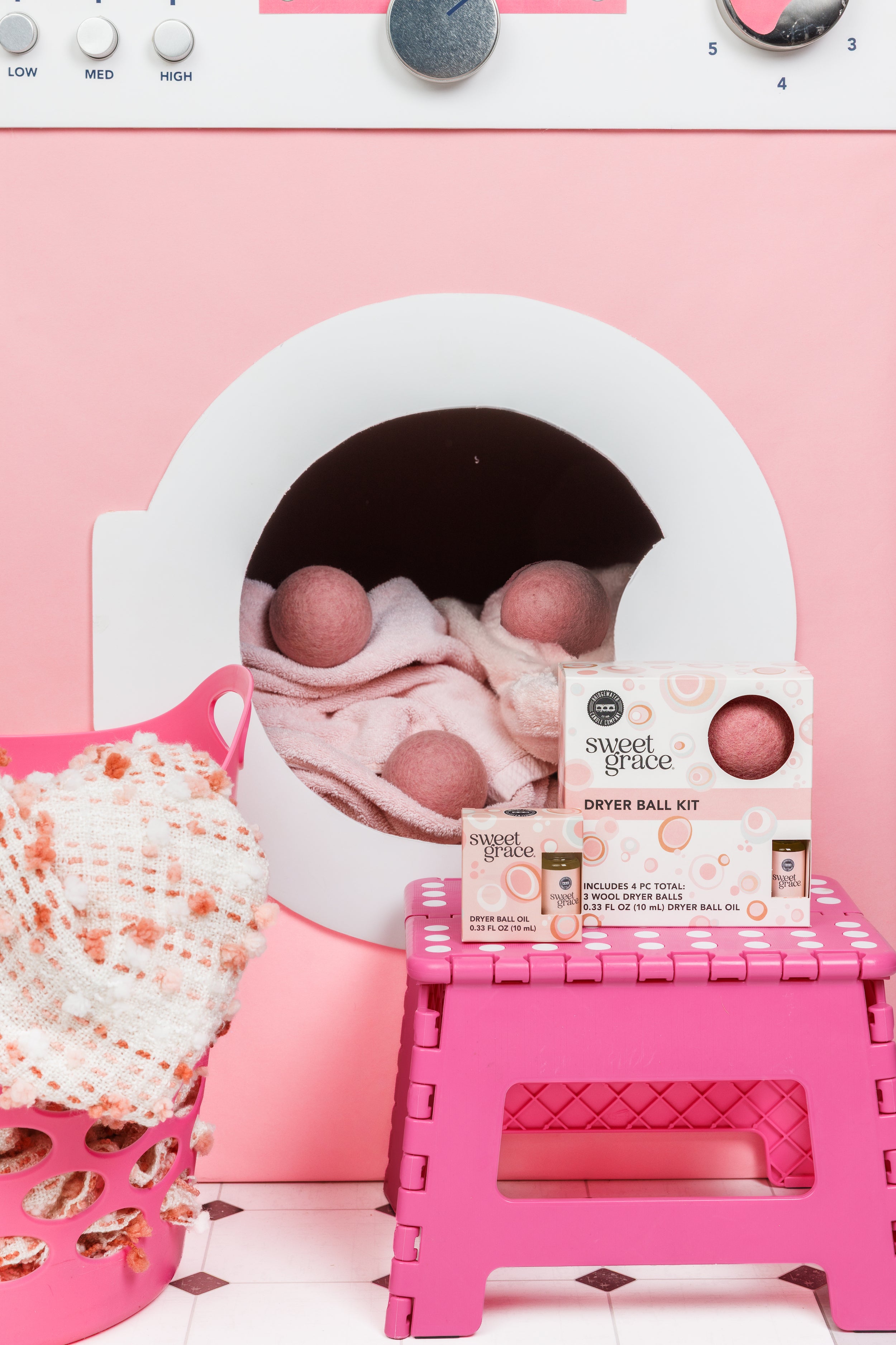 A pink-themed laundry setup with a dryer containing pink dryer balls, a basket with towels, a pink step stool, and a Sweet Grace dryer ball kit box in front.