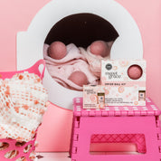 A cozy laundry scene features the Bridgewater Candle Sweet Grace Dryer Ball Kit, with wool dryer balls and kit box on a dryer, a pink stool, and a basket holding a soft pink and white blanket for an inviting, fragranced space.