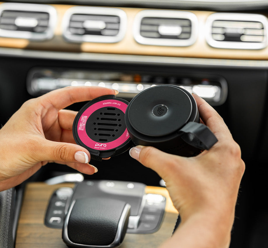 Person holding a Let's Celebrate pura car refill and pura car device inside the front of a car's interior.