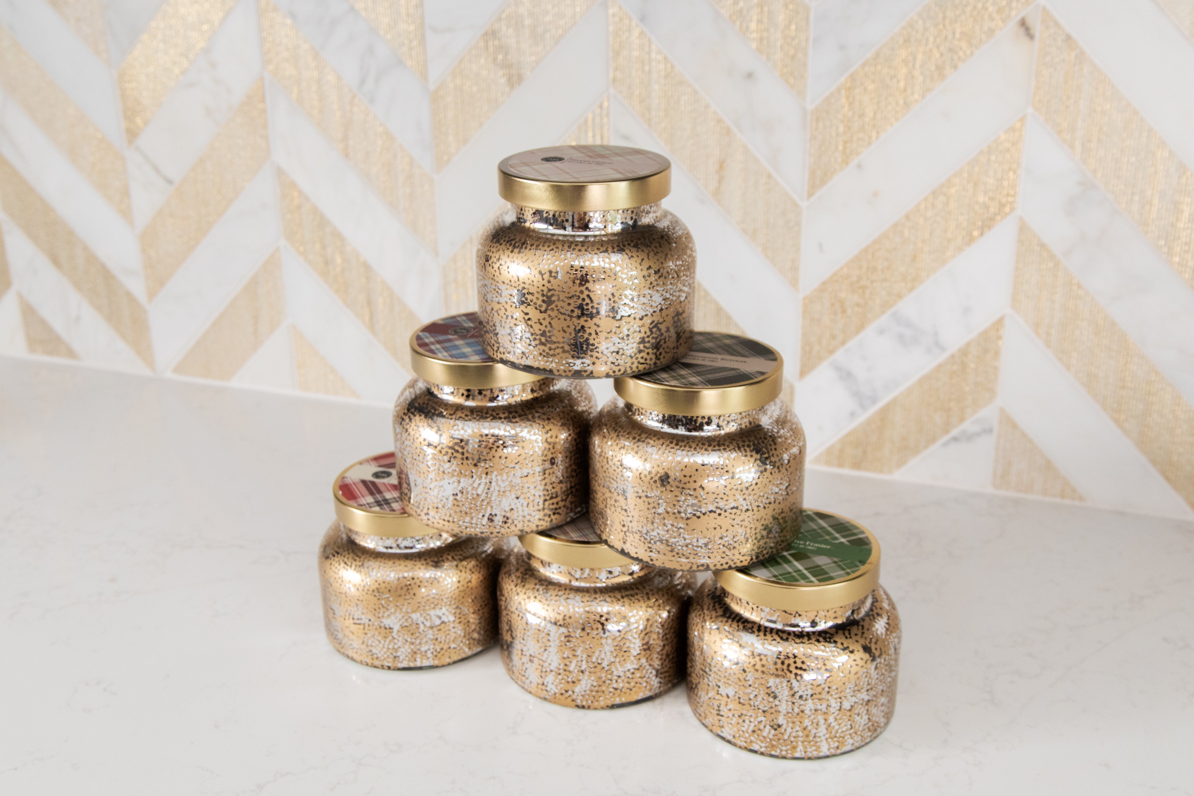Six Bridgewater Candle Company Christmas Bliss Holiday Mercury Jar Candles with gold lids are stacked pyramid-style on a white countertop before a gold-and-white chevron wall—ideal for adding festive cheer with their soy-blend wax.