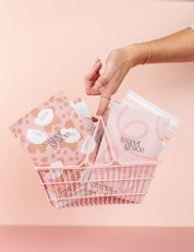 A hand holds a pink wire basket filled with Bridgewater Candle Companys Sweet Grace Modern Flower Sachets, featuring pastel colors and various patterns, against a soft pink background.