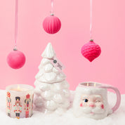 A Bridgewater Candle Sweet Grace Collection Candle #70, a white ceramic Christmas tree, and a winking Santa mug are arranged on faux snow, with pink ornaments above—all set against a pastel pink backdrop for festive decor.