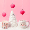 A white ceramic Christmas tree, the Bridgewater Candle Sweet Grace Holiday Nutcracker Candle, and a winking Santa mug rest on snow-like material. Three pink ornaments hang above, all against a pastel pink background for a whimsical holiday vibe.