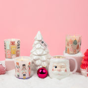 A festive centerpiece featuring the Bridgewater Candle Sweet Grace Holiday Tree Candle in decorative jars, a white ceramic Christmas tree, a winking Santa mug, pink ornaments, and faux snow—all set against a pink background for holiday cheer.
