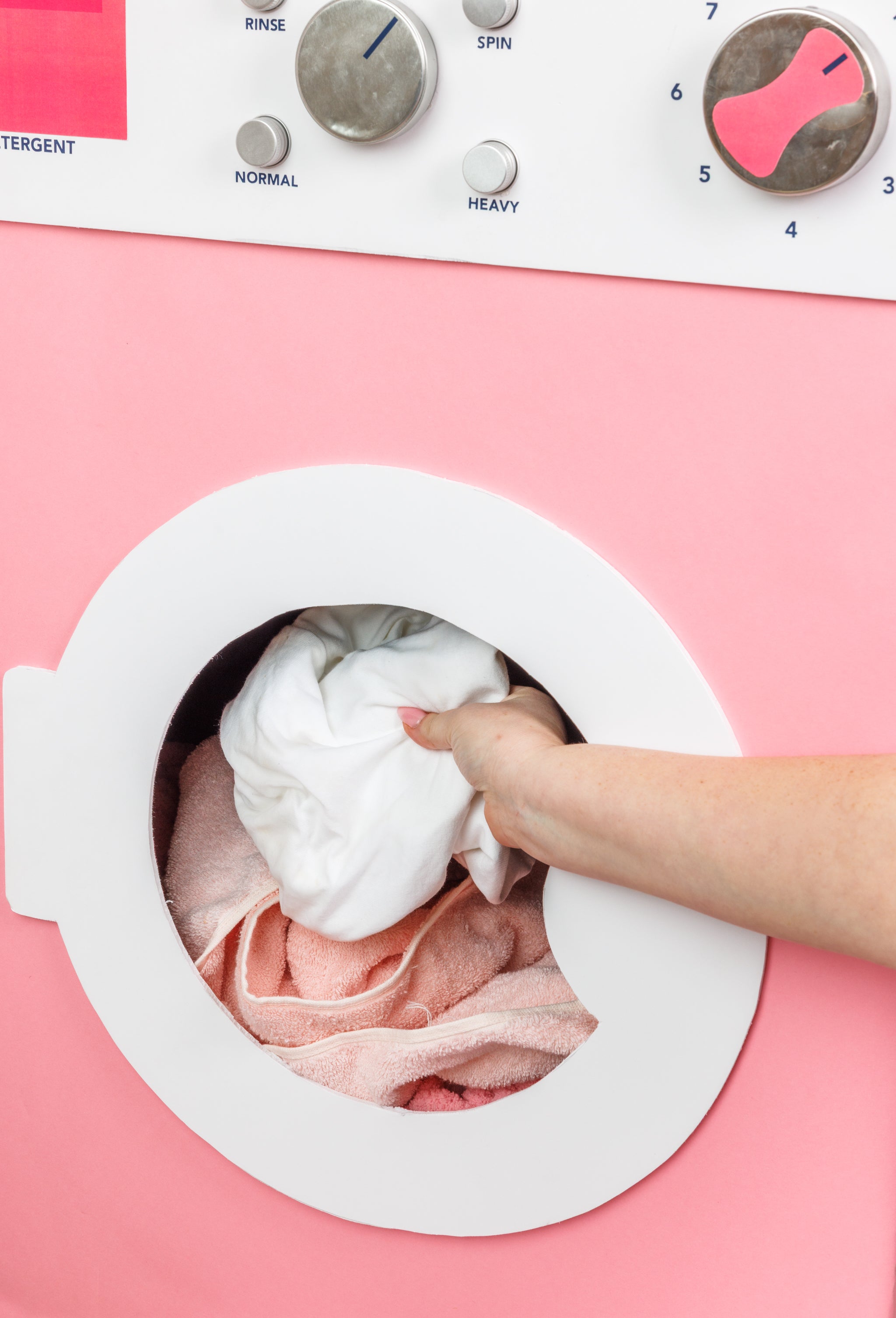 A persons hand places a white cloth into the drum of a front-loading washing machine with a pink exterior and control dials above the door.