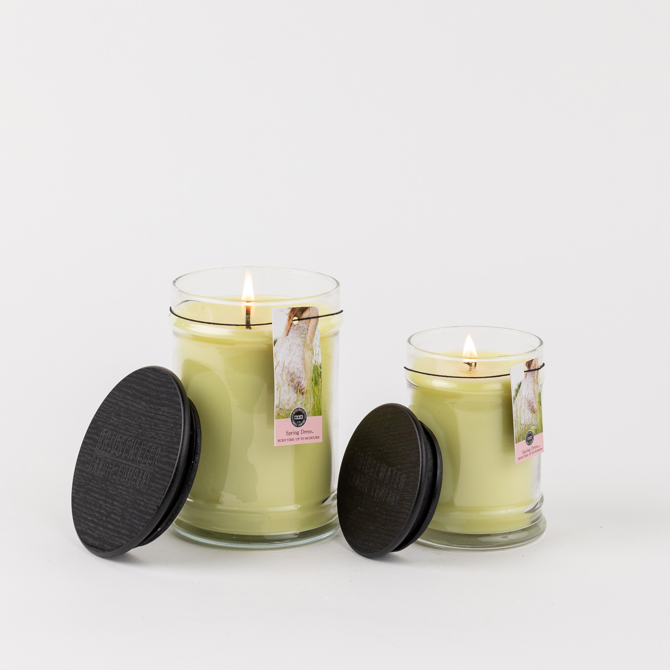 A large and a small lit yellow candle in glass jars with black lids sit on a white background. The small jar, the Spring Dress 8oz Jar Candle by Bridgewater Candle Company, offers a lovely inviting fragrance. Both display swan tags and text.