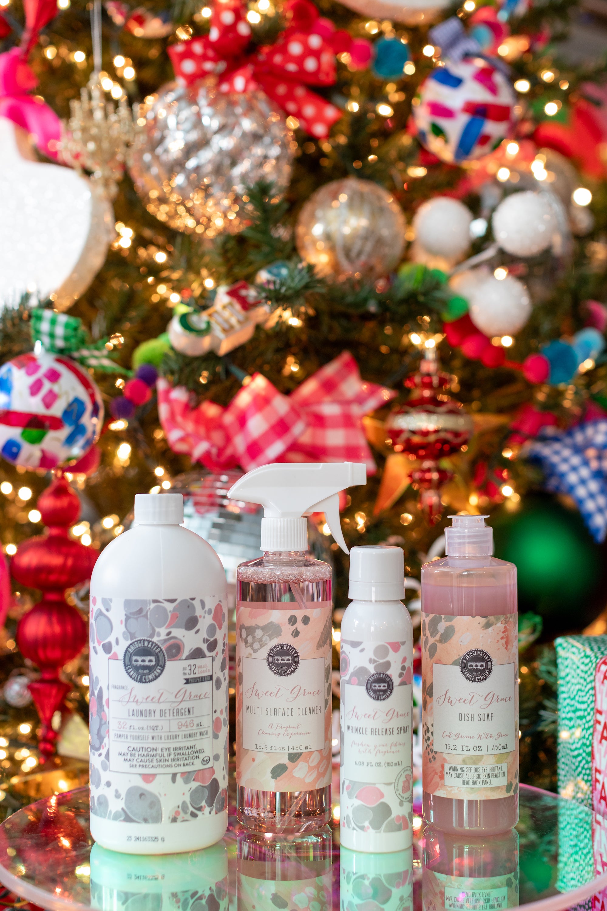 Four bottles of cleaning products are displayed on a table in front of a decorated Christmas tree with colorful ornaments and lights in the background.