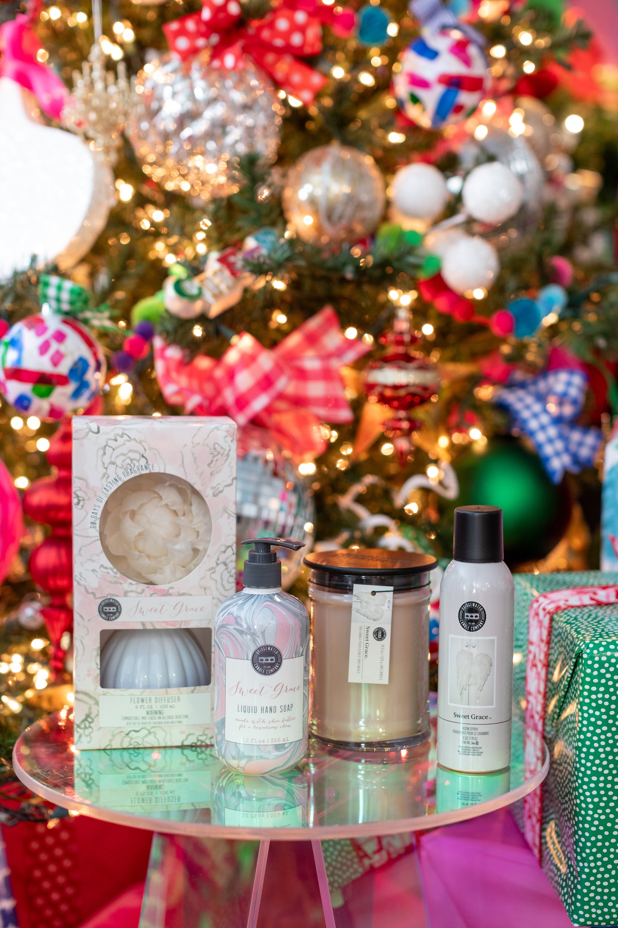 A glass table in front of a decorated Christmas tree holds a candle, a boxed truffle bath soap, a bottle of liquid hand soap, and a can of dry shampoo. Wrapped gifts are visible in the background.