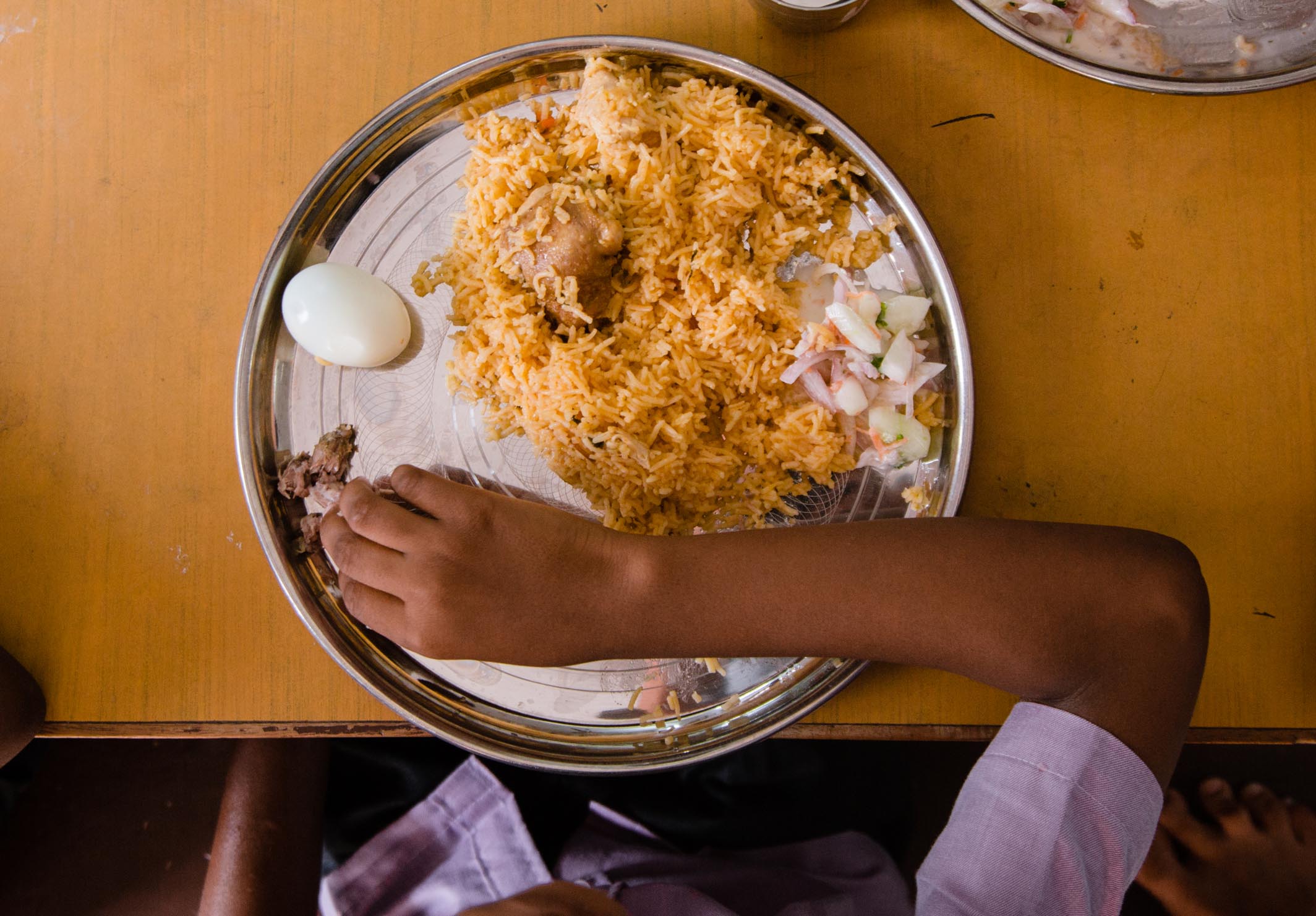 A child’s hand reaches for food on a metal plate with rice, a boiled egg, salad, and a piece of meat, placed on a wooden table. The child is wearing a light purple shirt.