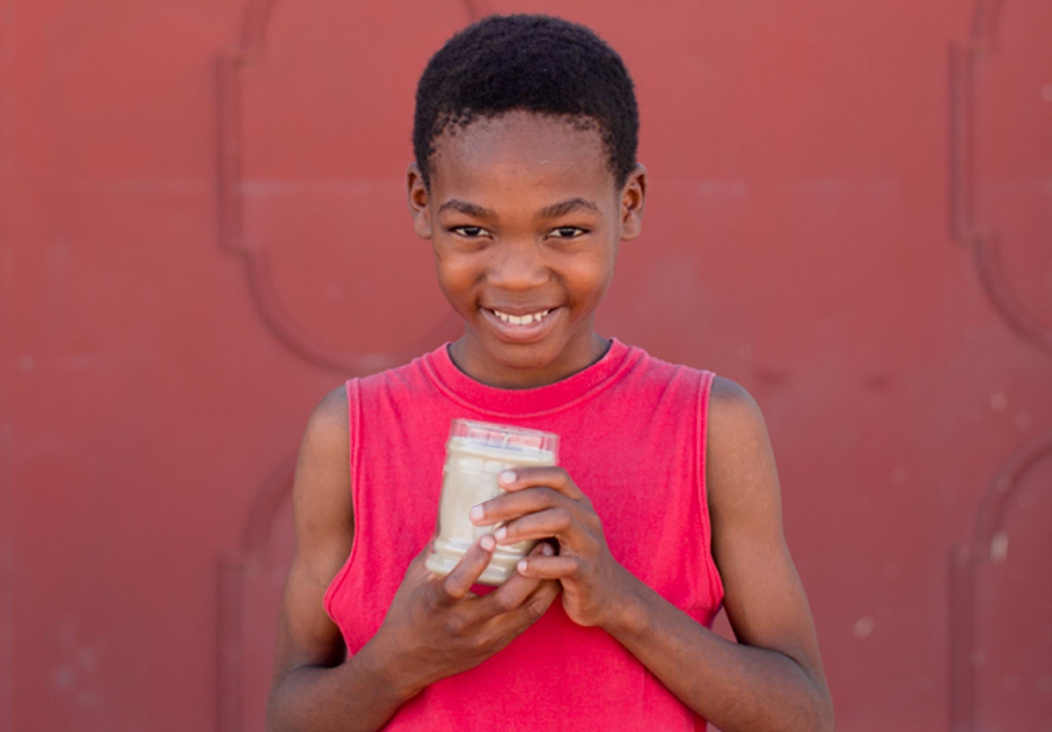A smiling child in a sleeveless red shirt holds a clear jar filled with coins and bills, standing in front of a reddish wall.