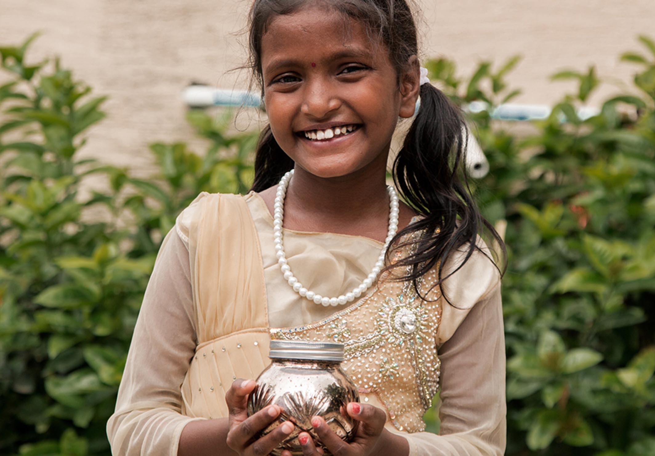 Smiling young girl in a beige dress and pearl necklace holding a silver jar, standing outdoors with green foliage in the background.
