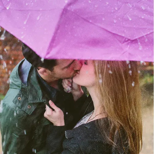 A couple kisses under a pink umbrella in the rain, with water droplets falling around them. The umbrella partially shields them from the rain.