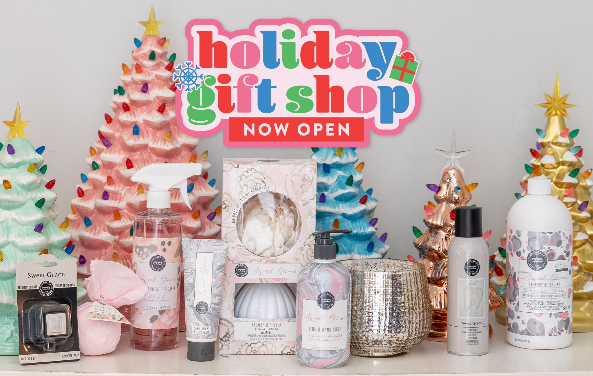 Festive display of candles, bath products, and sprays in front of pastel ceramic Christmas trees, with a colorful Holiday Gift Shop Now Open sign above.