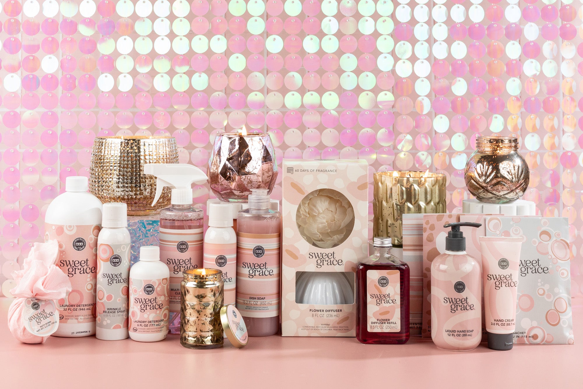 A collection of “Sweet Grace” scented products, including candles, sprays, lotions, soaps, and gift boxes, displayed on a pink surface with a shimmery, iridescent pink sequin backdrop.