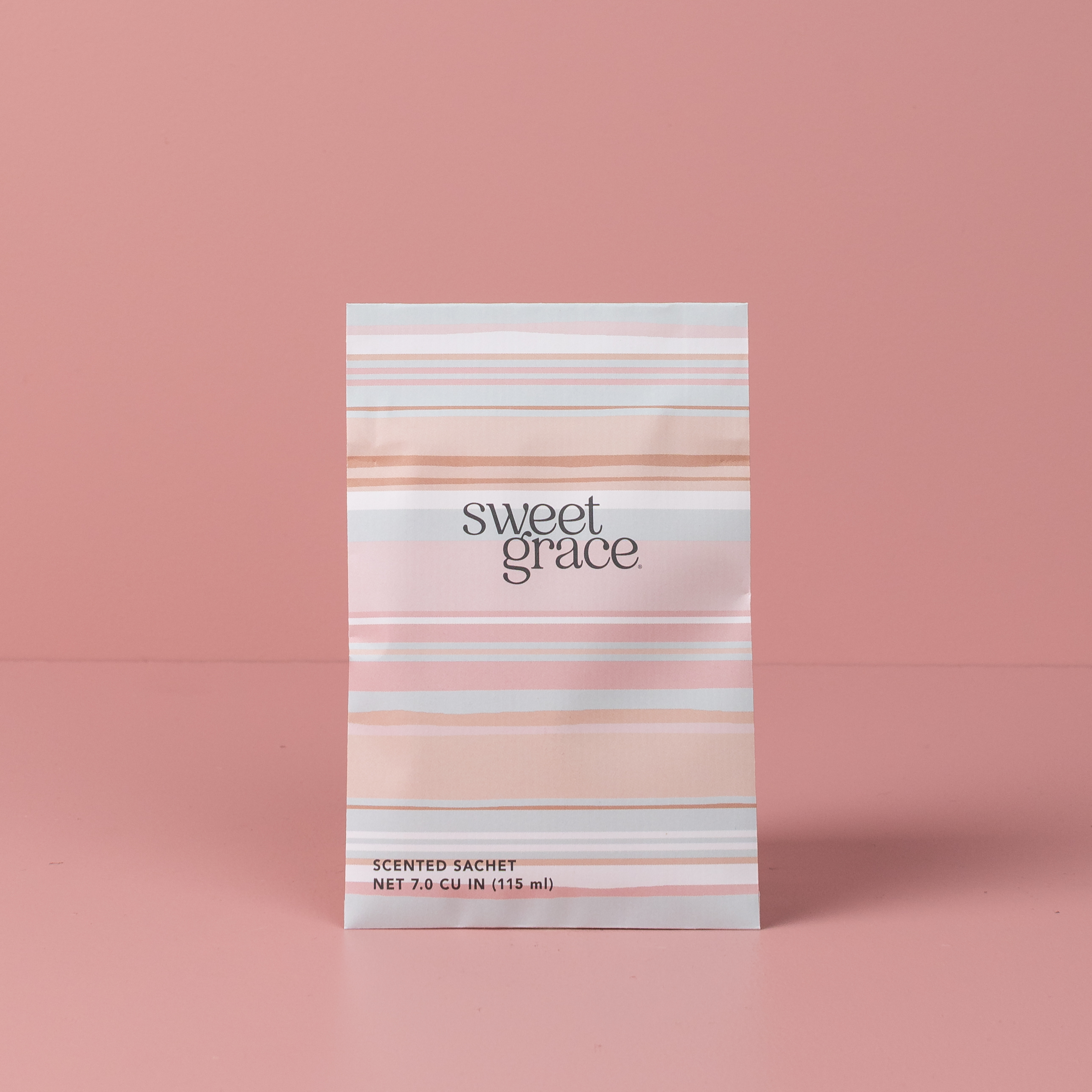 The Bridgewater Candle Company Sweet Grace Modern Stripe Sachet, infused with the signature Sweet Grace scent, stands upright on a pink surface and background. The sachet features pink and peach stripes with the product name displayed on the front.