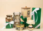 A curated gift set featuring Bridgewater Candle Companys Afternoon Retreat Large Decorative Candle, soy-blend wax, jars, and green gift boxes with white ribbons—all elegantly displayed beside a cream artificial flower in a gold vase.