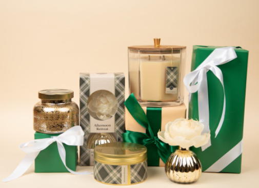 A collection of home decor, featuring a Bridgewater Candle Company Afternoon Retreat Mercury Jar Candle, a gold jar, and two green gift boxes with white ribbons, is displayed alongside a cream flower in a gold vase against a beige background.
