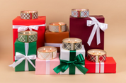 A group of colorful gift boxes with ribbons are topped with Bridgewater Candle Company Comfort & Joy Holiday 3-Wick Tin Candles. The background is plain light brown.