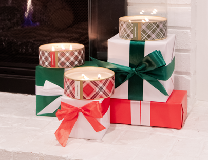 Three wrapped gift boxes with green and red ribbons are stacked by a fireplace, each topped with a lit Festive Frasier Holiday 3-Wick Tin Candle by Bridgewater Candle Company, made from soy-blend wax.