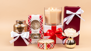 A festive scene features Bridgewater Candle Company's Comfort & Joy Holiday 3-Wick Tin Candle, surrounded by gift-wrapped boxes, candles, and jars with white and red ribbons. A white flower accents a gold-striped vase against a beige background.