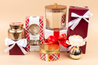 A festive display features the Bridgewater Candle Company Comfort & Joy Holiday Large Decorative Candle with soy-blend wax and premium fragrance, a flower diffuser, and gift boxes in gold, red, and plaid on a beige background.