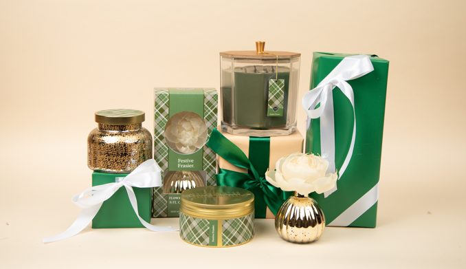 A selection of green and gold holiday gifts featuring the Bridgewater Candle Company Festive Frasier Holiday Mercury Jar Candle, plus a decorative flower and wrapped presents with white ribbons, all set on a beige background.