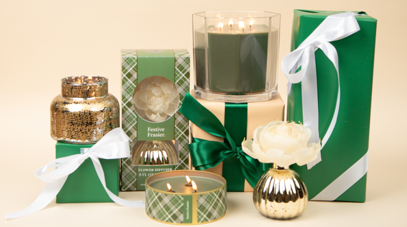 A set of green and gold gift boxes with white ribbons, plus Bridgewater Candle Company's Festive Frasier Holiday 3-Wick Tin Candle, arranged on a beige background.