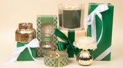 A set of green and gold gift boxes with white ribbons, plus Bridgewater Candle Company's Festive Frasier Holiday 3-Wick Tin Candle, arranged on a beige background.
