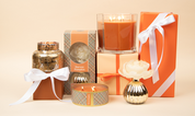 A display of Bridgewater Candle Company's Harvest Pumpkin Holiday Large Decorative Candle, set in glass and metallic holders, surrounded by orange and cream gift boxes with white ribbons against a beige background.