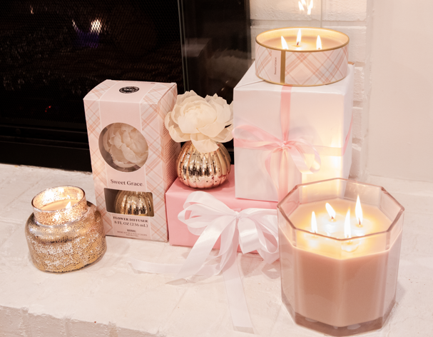 A Sweet Grace Large Decorative Candle by Bridgewater Candle Company, gift boxes with white and pink ribbons, a flower in a gold vase, and a floral-scented diffuser decorate the white brick hearth before the fireplace.