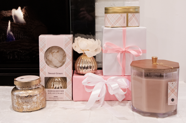 The Sweet Grace Large Decorative Candle by Bridgewater Candle Company is arranged with pink and gold-wrapped gifts and a flower diffuser on a white surface before a lit fireplace, creating a cozy, elegant scene.