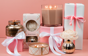 Pink and white gift-wrapped boxes with ribbons, candles—including the Bridgewater Candle Company Sweet Grace 3-Wick Tin Candle—and a flower-shaped diffuser are arranged against a pink background.