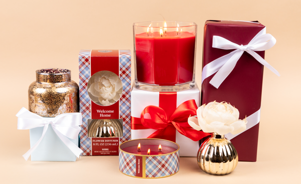 The Welcome Home 3-Wick Tin Candle by Bridgewater Candle Company is presented in festive red, gold, white, and plaid packaging with white ribbon bows and displayed on a peach background.
