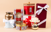 The Bridgewater Candle Company’s Welcome Home Large Decorative Candle features a soy-blend wax in an elegant gold jar, paired with wrapped gift boxes, a plaid tin, and floral accents—perfect for stunning home decor.