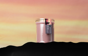 A Bridgewater Candle Company Hello Night 18oz Jar Candle in a glass jar with a gold lid sits against a yellow and pink sunset. Crafted from soy-blend wax, it features a tag and is framed by a dark, wavy silhouette.