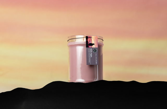 A Bridgewater Candle Company Hello Night 18oz Jar Candle in a glass jar with a gold lid sits against a yellow and pink sunset. Crafted from soy-blend wax, it features a tag and is framed by a dark, wavy silhouette.