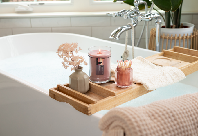 A bathtub with a wooden tray holds a lit Hello Night 18oz Jar Candle by Bridgewater Candle Company (soy-blend wax), a small vase of dried flowers, cotton swabs in a glass jar, and a rolled towel. Natural light and green plants surround the scene.