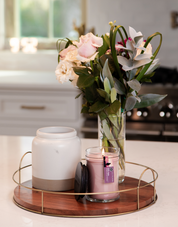 A vase of pink and white flowers, a white ceramic jar, and a lit Hello Night 18oz Jar Candle by Bridgewater Candle Company sit on a wooden tray with brass handles on a kitchen countertop.