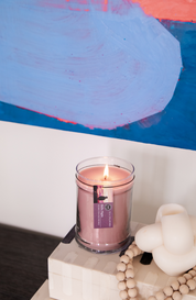 A lit Hello Night 18oz Jar Candle by Bridgewater Candle Company sits on stacked books beside a white knotted decor and wooden beads, with a colorful abstract painting in the background.