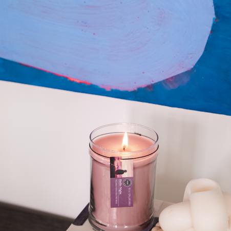 A lit Hello Night 18oz Jar Candle by Bridgewater Candle Company sits on stacked books beside a white knotted decor and wooden beads, with a colorful abstract painting in the background.