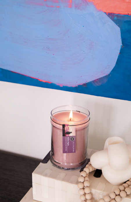 A lit Hello Night 18oz Jar Candle by Bridgewater Candle Company sits on stacked books beside a white knotted decor and wooden beads, with a colorful abstract painting in the background.