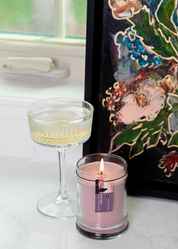 A glass of sparkling wine, a lit Bridgewater Candle Company Hello Night 8oz Jar Candle, and a colorful floral painting are displayed on a white marble surface near a window.