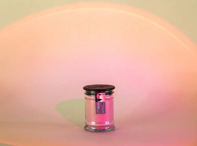 A Bridgewater Candle Company Hello Night 8oz Jar Candle with a black lid and tag sits on a smooth surface, softly lit by pink-orange gradient light. Made from soy-blend wax for a clean, long-lasting burn.
