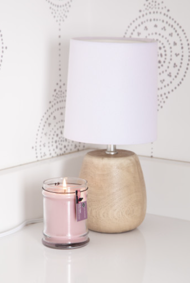 A lit Bridgewater Candle Company Hello Night 8oz Jar Candle sits in a glass jar beside a small table lamp with a wooden base and white shade, all arranged on a white surface against a white patterned wall.